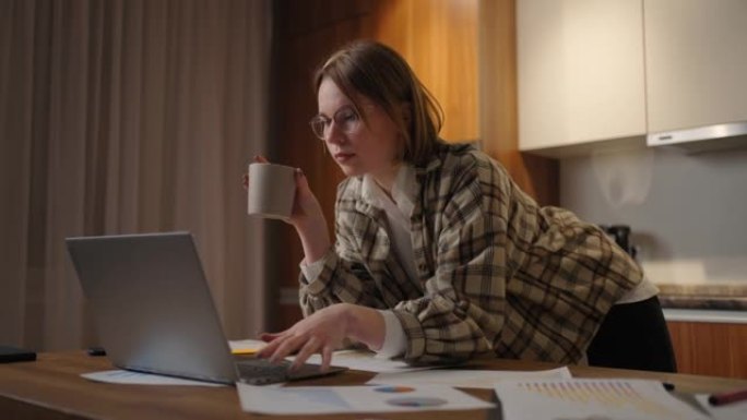 美丽的年轻女子坐在客厅喝咖啡时在笔记本电脑上工作。在笔记本电脑上工作的快乐休闲美女。在家使用笔记本电