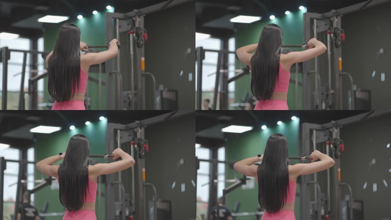 一个穿着粉红色西装的黑发女人将跨界训练器中的绳子拉到她的胸前。在教练中进行背部锻炼。专业女性教练
