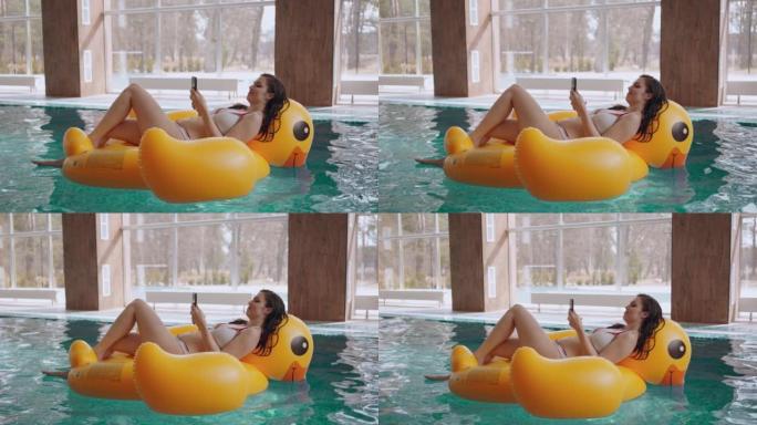 穿着比基尼的苗条女人躺在游泳池里的黄色充气游泳圈上，用手机