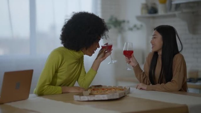 轻松大笑的非洲裔美国人和亚裔妇女坐在厨房里，酒杯和比萨饼在说话。积极快乐的女性朋友周末在室内休息。友