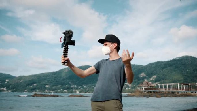 一个男性视频记录器在海滩附近戴着口罩与手持相机交谈的特写镜头