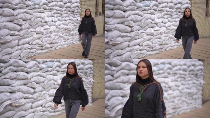 宽镜头严肃自信的年轻女子穿着刺绣衬衫，在乌克兰城市通过保护沙袋墙离开慢镜头。美丽的女士走在街上环顾四