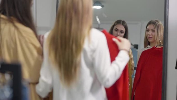 体贴苗条的女人在商店镜子里倒影，朋友带着衣架说话。两个积极的朋友换毛衣在室内购物。决策与消费主义概念
