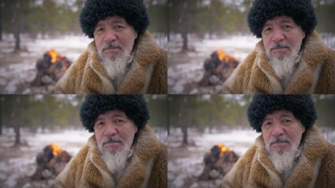 特写肖像严肃的土著高级大胡子男子看着坐在右边的相机，背景着火。老亚洲蒙古人在森林里慢动作摆姿势。
