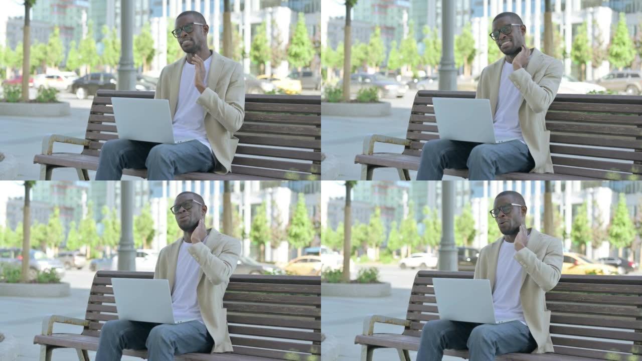 坐在户外长凳上时使用笔记本电脑颈部疼痛的非洲男子坐在户外长凳上时使用笔记本电脑颈部疼痛的非洲裔美国人