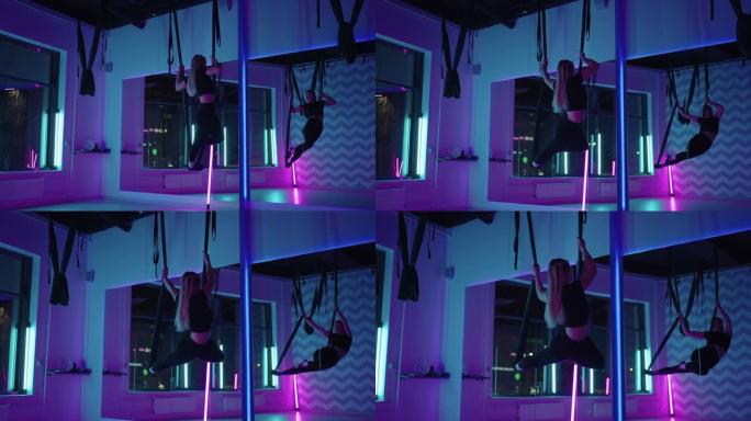 一名妇女在霓虹灯下的吊床上空中做运动瑜伽，以慢动作进行优美的练习和动作。