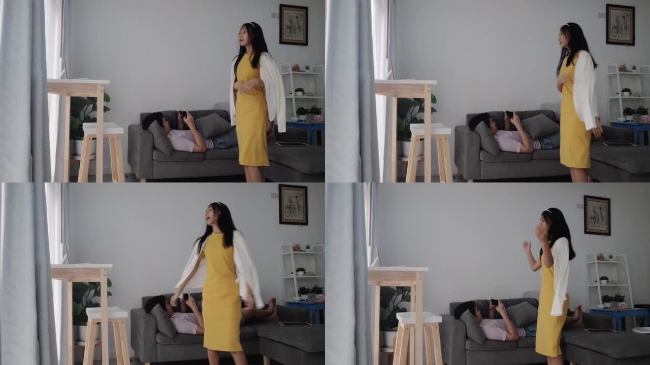亚洲女孩在躺在沙发上在家玩手机游戏的哥哥附近制作病毒视频和跳舞，这是新一代生活方式的概念。