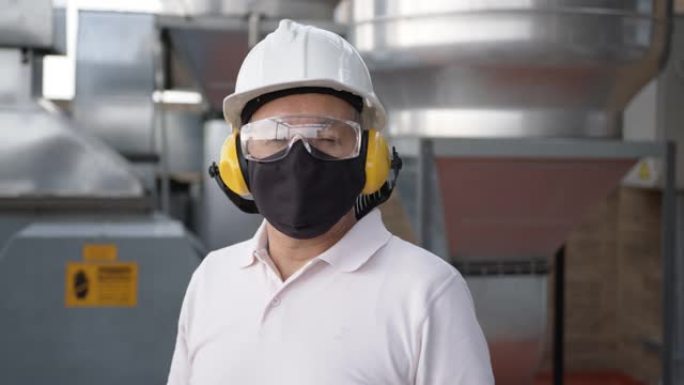 在工厂面对摄像机的男主管的特写镜头，戴着护耳器，护目镜，头盔和防护面罩