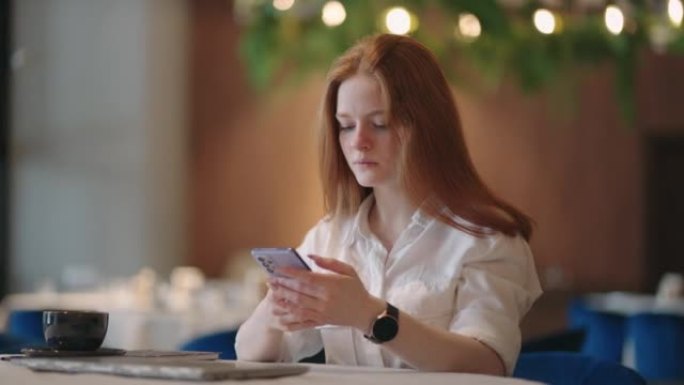 红发女人在家工作时使用笔记本电脑，同时在手机上阅读短信。女人用电话。认真迷人的女人在家里使用笔记本电