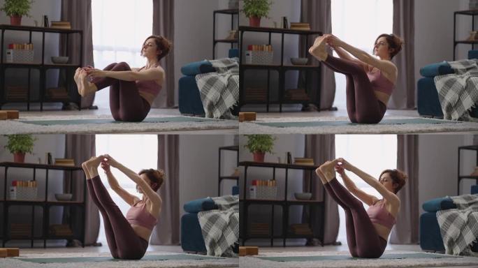 运动的女人正在家里练习瑜伽，坐在客厅里，在空中保持双腿，拉紧肌肉