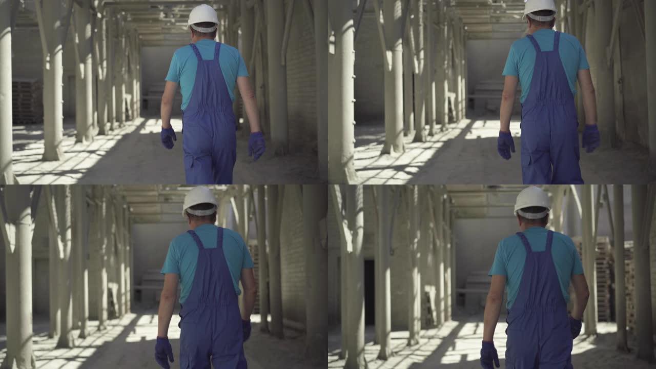 穿着制服和头盔的成年白人男子在阳光下走在建筑工地上的后视图。摄像机跟随男性工人在阳光明媚的夏日漫步在
