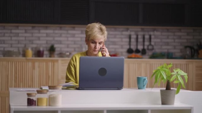 老年女士正在远程工作，坐在家里的厨房里，用手机交流，用笔记本电脑发送电子邮件