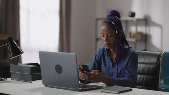 年轻的黑人妇女坐在家里的工作场所，使用智能手机，在社交网络中观看新闻录像带，玩游戏