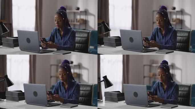 年轻的黑人妇女坐在家里的工作场所，使用智能手机，在社交网络中观看新闻录像带，玩游戏