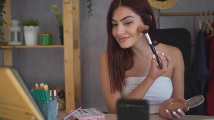 美丽微笑的女红头发展示如何用刷子在脸上涂抹化妆粉