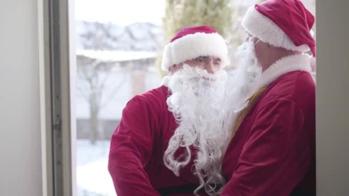 两个大肚子的圣诞老人进入房子的侧视图。在圣诞节或新年前夕，穿着红色圣诞老人服装的年轻人和高级男子带着