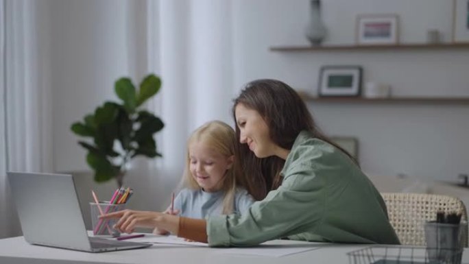 漂亮的母亲帮助女儿坐在家里的桌子旁做作业。女儿在家和妈妈一起上在线课。用电脑远程学习的孩子。