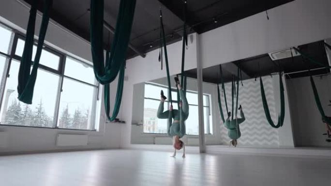 一个年轻漂亮的女人在空中瑜伽的吊床上练习。下午，一名妇女在大厅的镜子前独自表演伸展运动