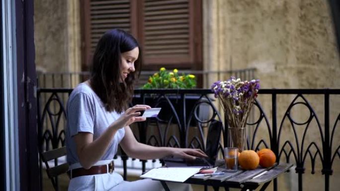 年轻女子坐在阳台上用笔记本电脑用信用卡在线购物