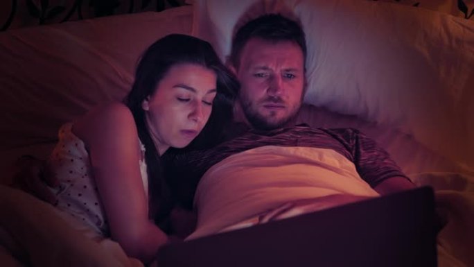 年轻夫妇在床上看恐怖电影。使用笔记本电脑