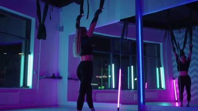 一名妇女在霓虹灯下的吊床上空中做运动瑜伽，以慢动作进行优美的练习和动作。