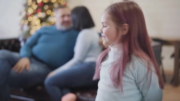 开朗的高加索女儿看着相机，跑回坐在背景沙发上的快乐父母。迷人的女孩在家享受圣诞假期庆祝。机架聚焦。