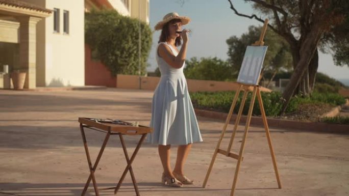 自信的创意白人妇女，穿着优雅的连衣裙和草帽，在阳光下的画架上喝着红酒欣赏照片。苗条优雅的艺术家站在阳