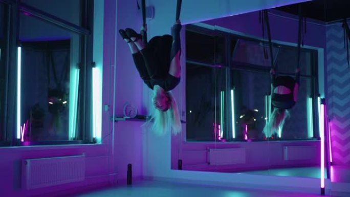 一个女人在霓虹灯下做瑜伽并在悬吊的吊床上伸展。年轻漂亮苗条的身体健身女孩在健身房练习飞行瑜伽。
