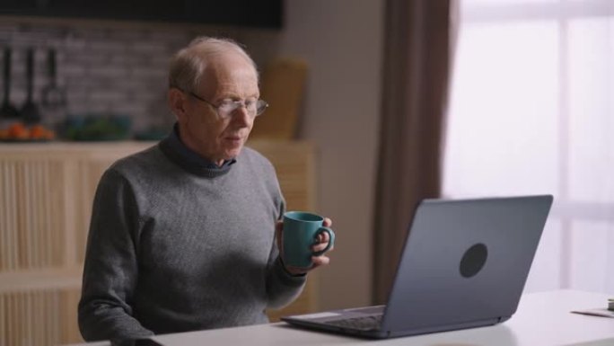 祖父正在通过视频通话进行交流，独自一人坐在家里的厨房里，向笔记本电脑的网络摄像头挥手，欢迎对话者