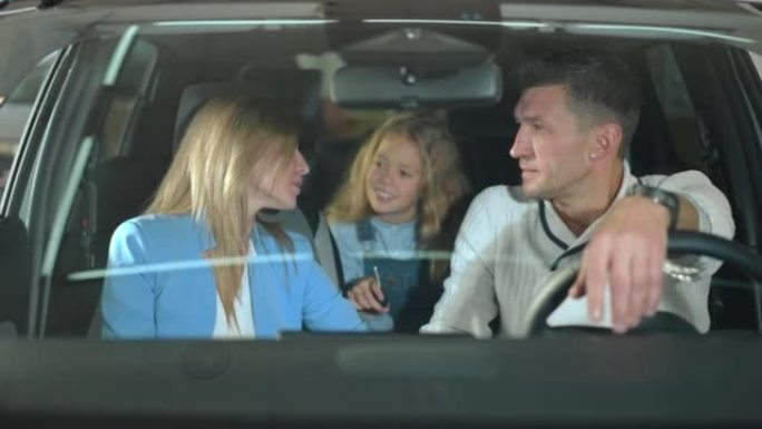 快乐的父母和坐在经销商的新车里的女儿交谈。积极的白人男人和女人在陈列室里和女孩一起选择汽车。家庭和消