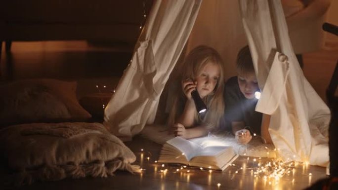 幸福的家庭。两个孩子女孩和男孩正在帐篷里看书。兄弟姐妹手中的手电筒。帐篷里有书的幸福家庭。孩子们坚持