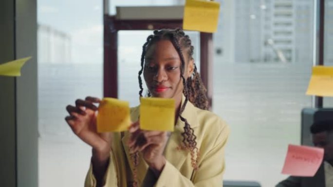 非裔美国商业女性在玻璃板上的便利贴上写任务和创意，女性公司领导策划贴贴便利贴上的项目