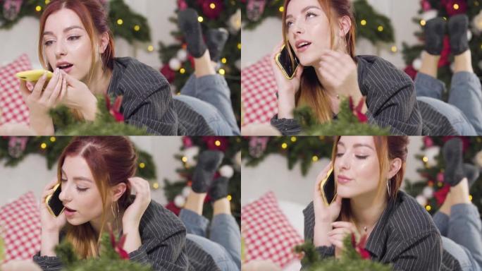 年轻的红发女人在除夕夜用虚拟助手打电话给某人。迷人美丽的高加索女士的肖像祝贺朋友躺在床上的圣诞节。假