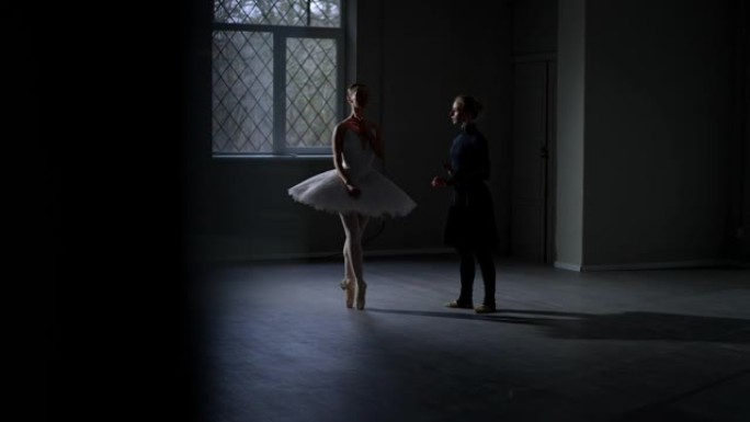 穿着白色短裙的优雅芭蕾舞演员在黑暗中排练，编舞穿着黑色连衣裙。两个漂亮的高加索年轻女子在工作室室内跳