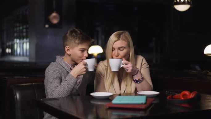 放松的白人妇女和男孩坐在咖啡馆的餐桌旁，闻到慢动作喝热美味的咖啡的味道。中景肖像快乐母子在餐厅享受家