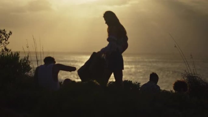 一群不同的朋友坐在绿草如茵的悬崖上，看着海上的日落。一起露营