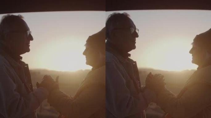 两个老年人在公路旅行中露营。站在大型汽车后备箱旁边互相暖手垂直视频