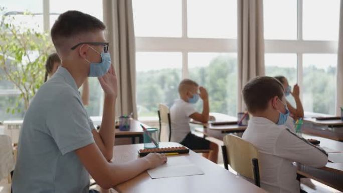 学校的孩子们坐在教室里，举手慢动作地戴着防护面具回答老师的问题