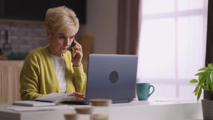 老妇人在家工作，用手机聊天，输入笔记本电脑，家庭办公室和老年人的远程工作模式