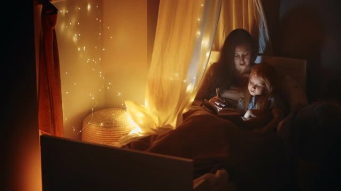 可爱的小女孩和她的母亲在圣诞节期间在黑暗的卧室里一起看书