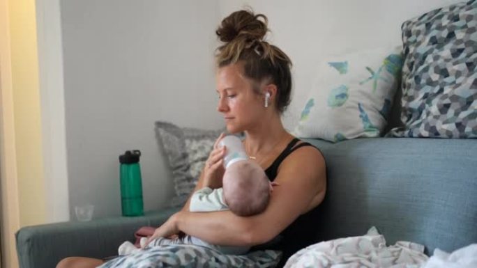母亲在家中的沙发上喂养婴儿，戴着耳机观看智能手机屏幕