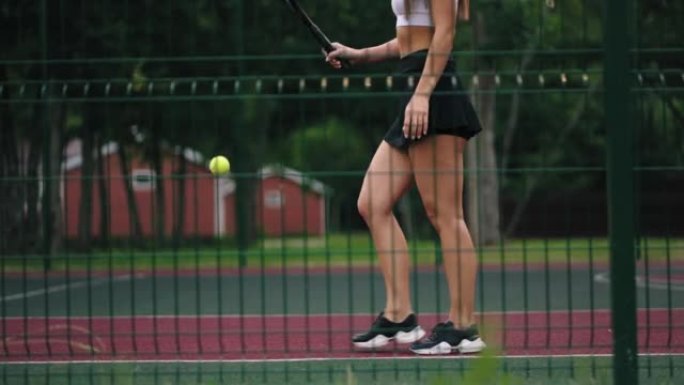 运动的年轻女子正在球场上练习网球，走路和弹跳球，修长的双腿