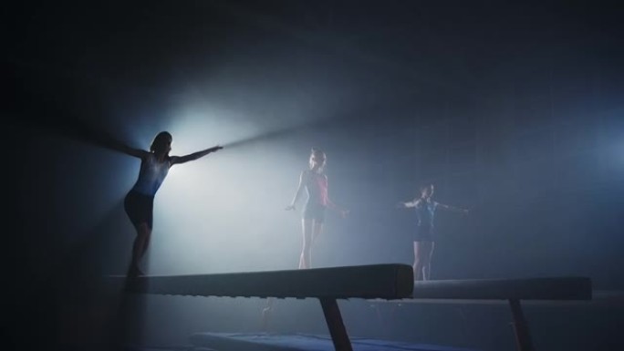 三名女体操运动员在艺术体操运动员团队的黑暗体操馆的平衡木上表演