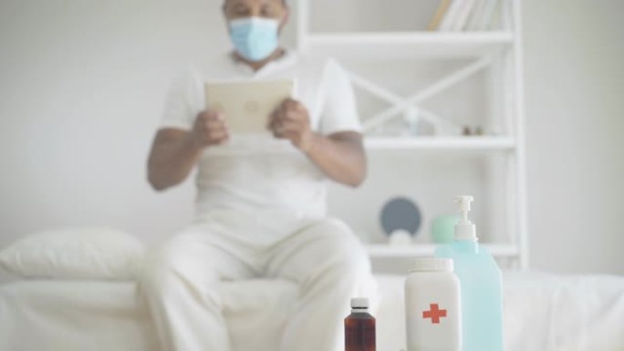 背景下使用平板电脑的消毒剂和药丸与模糊的非洲裔美国人的特写镜头。生病的男性患者戴着口罩坐在医院病房的
