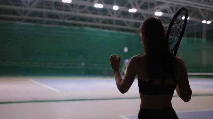 年轻的黑发女子在室内的网球场上行走，投掷球并接球，准备发球。