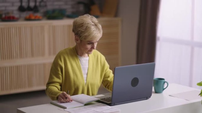 老妇人正在网上学习，坐在家里的厨房里拿着笔记本电脑，在网上寻找信息，在笔记本上做笔记