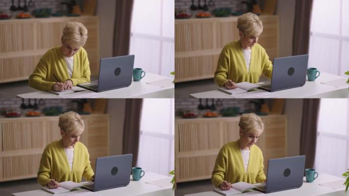 老妇人正在网上学习，坐在家里的厨房里拿着笔记本电脑，在网上寻找信息，在笔记本上做笔记