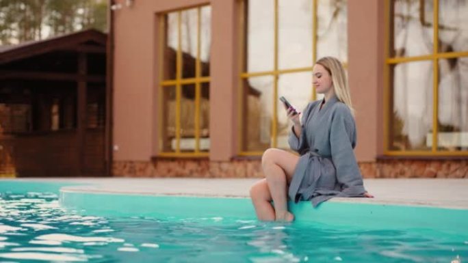 漂亮女人坐在室外热水池的边缘，休息，用现代智能手机上网