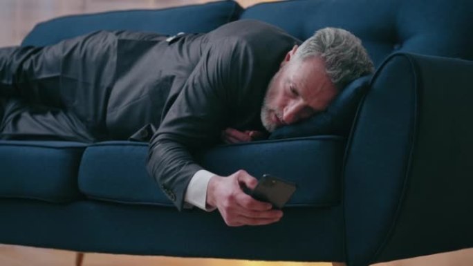 疲惫的商人躺在沙发上，通过电话阅读有关公司破产的新闻