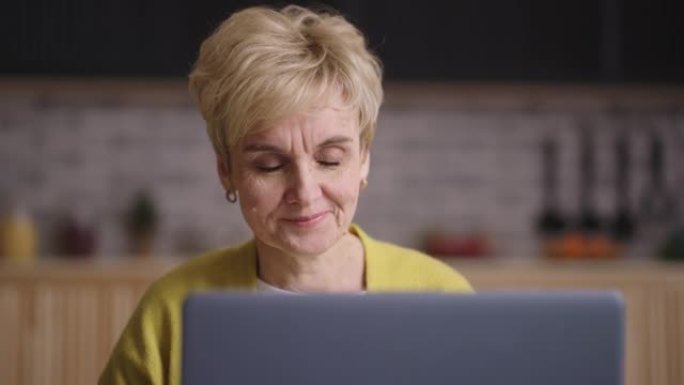 退休妇女正在笔记本电脑上通过视频聊天聊天，坐在家里的厨房里，脸上满是皱纹的微笑女士的肖像